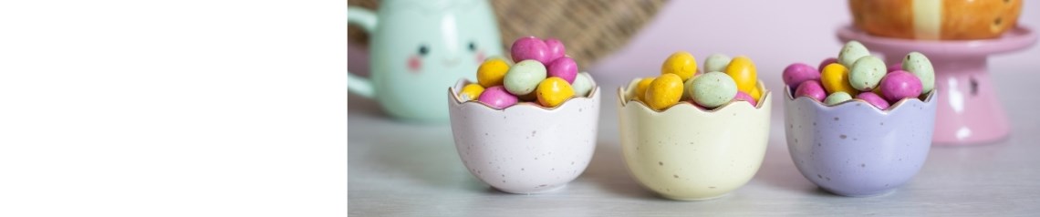 En påskkollektion för hemmet med pastellfärgade muggar, formade oljelampor och vårdekorationer med leende ägg och motiv av hot cross buns.